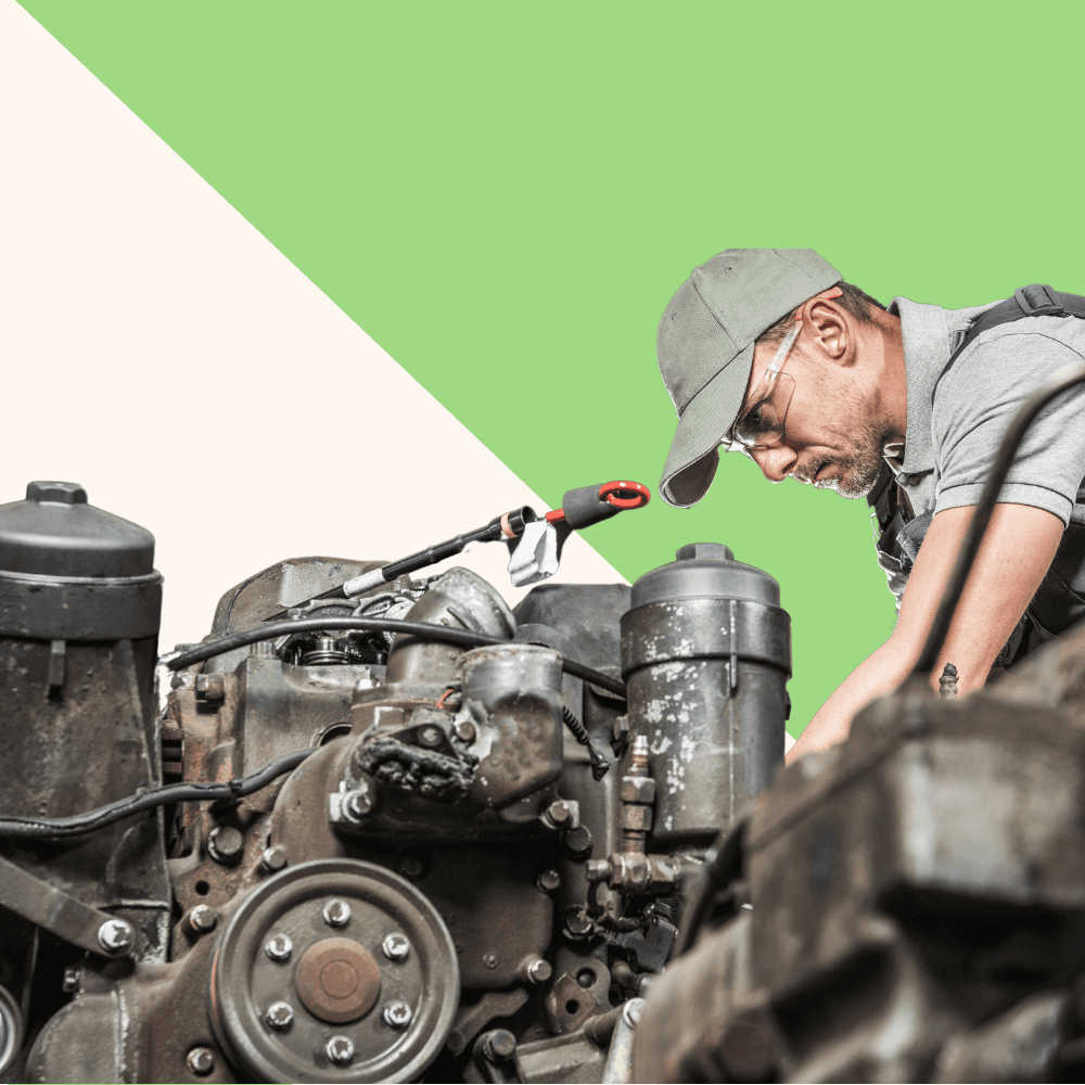Mechanic wearing cap and glasses works intently on engine. Background features green and cream geometric design. Concentrated expression. Industrial setting.