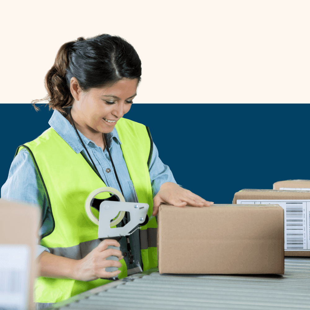 A woman in a high-visibility vest smiles while sealing a cardboard box with tape at a conveyor belt. The setting is a warehouse, conveying efficiency and focus.